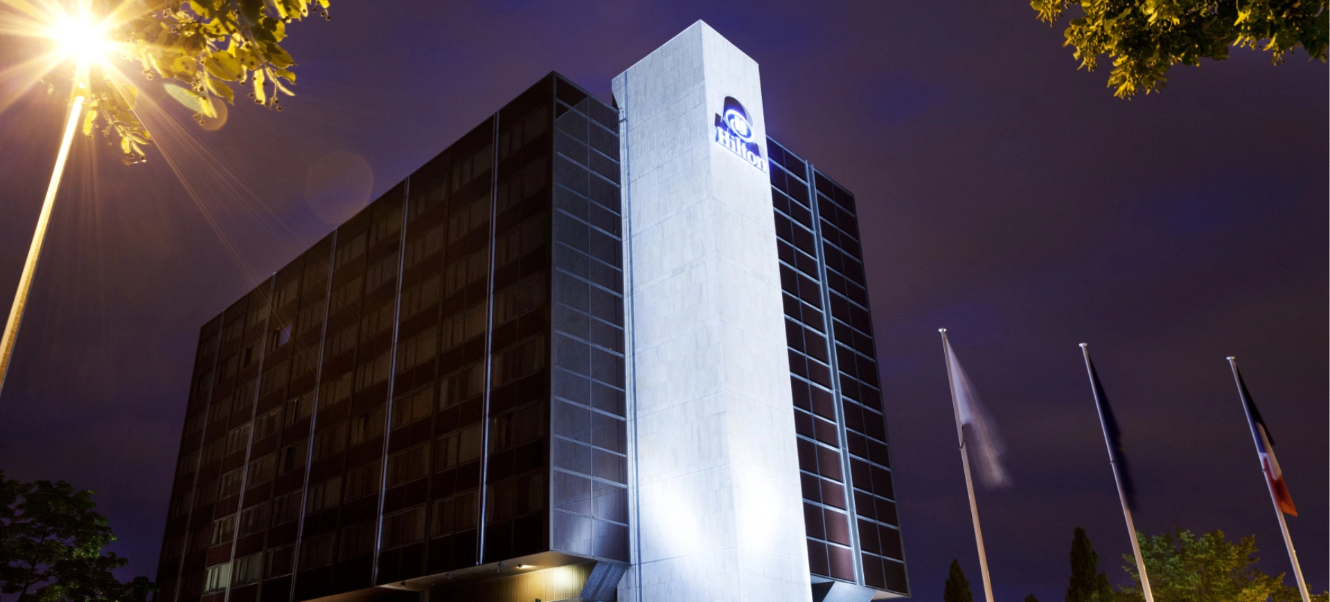 Vue nocturne d’un hôtel moderne éclairé, avec des drapeaux flottant devant l’entrée.