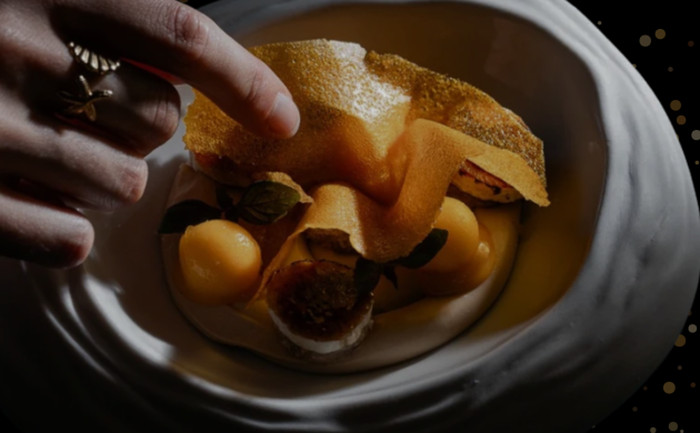 Dessert gastronomique raffiné décoré de feuilles et billes fruitées, servi dans une assiette élégante, avec une main en train de découvrir le plat.